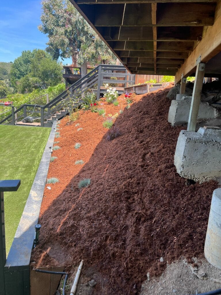 A sloped garden bed covered with brown mulch sits under a wooden deck, with small plants spaced throughout and retaining walls on either side.