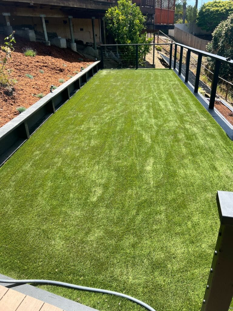 A rectangular area of artificial grass bordered by black railings and garden beds, situated in a backyard with sunlight casting shadows.