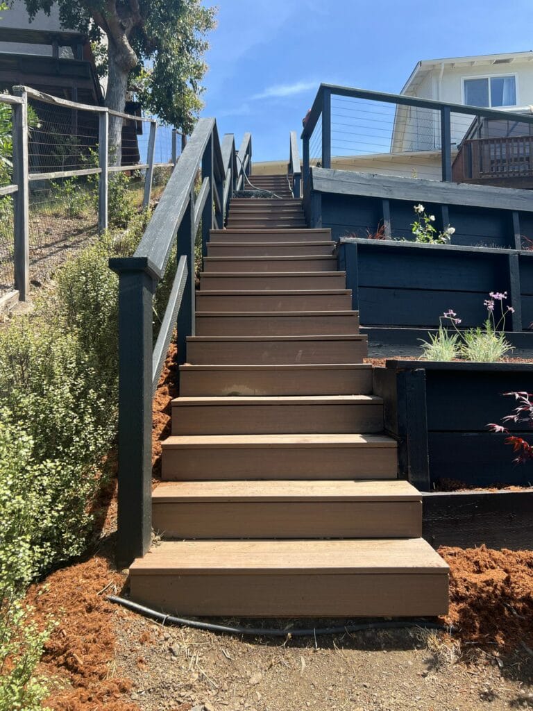 A wooden outdoor staircase with black railings leads uphill between garden beds, ending at a deck near a white house under a blue sky.