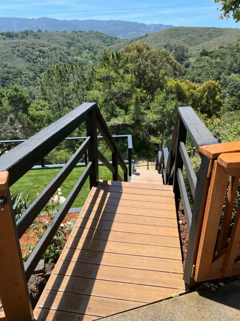 Wooden stairs with black railings descend toward a green lawn and forested hills under a clear blue sky.